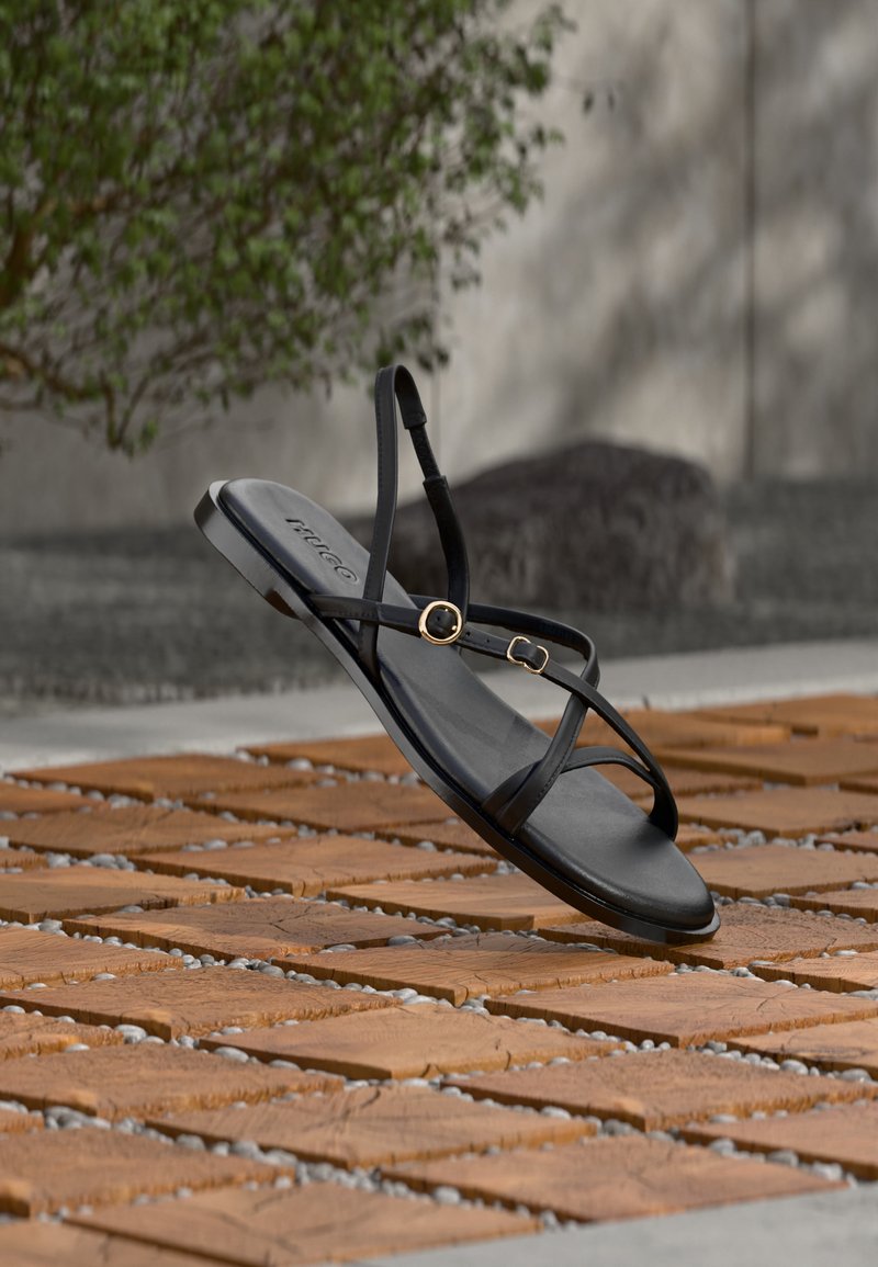 Μαύρο σανδάλι με λουράκια και επίπεδη σόλα, με χρυσά αγκράφες, που αιωρείται πάνω από ξύλινο πλακάκι σε εξωτερικό χώρο κοντά σε τοίχο και φυλλωσιά.