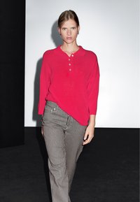 Maglione polo rosso con colletto e dettagli con bottoni, abbinato a pantaloni a gamba larga a righe bianche e nere, esposto su uno sfondo neutro.