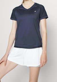 Námořnický sportovní top s krátkými rukávy a síťovanými akcenty, s výstřihem do V a rovnými lemy, v kombinaci s bílými sportovními šortkami.