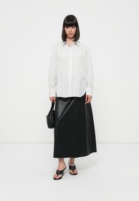 Camicia a righe bianche e grigie abbinata a una gonna maxi nera in pelle, sandali con tacco neri e una piccola borsa nera.