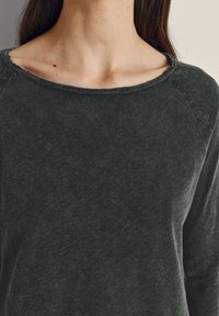 Nainen, joka pukeutuu tummanharmaaseen pyöreäkauluspaitaan, näkyvillä ovat solisluut ja pitkät ruskeat hiukset, yksinkertaista taustaa vasten.