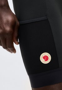 Κοντινό πλάνο σκούρου δέρματος χεριού δίπλα σε μαύρο ποδηλατικό σορτς με δίχτυ τσέπης και στρογγυλό κόκκινο λογότυπο αλεπούς.