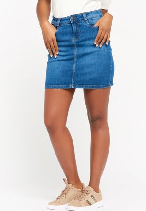 Frau trägt einen blauen Denim-Minirock, weißes Oberteil und beige Sneakers, steht mit Händen auf den Hüften vor weißem Hintergrund.