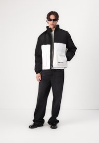 Black and white insulated jacket with a high collar, front zipper, and two large pockets; worn with loose black jeans and black shoes.