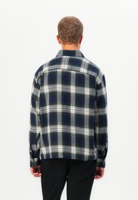 Man facing away wearing a black and white plaid long-sleeve shirt with black pants against a plain white background.