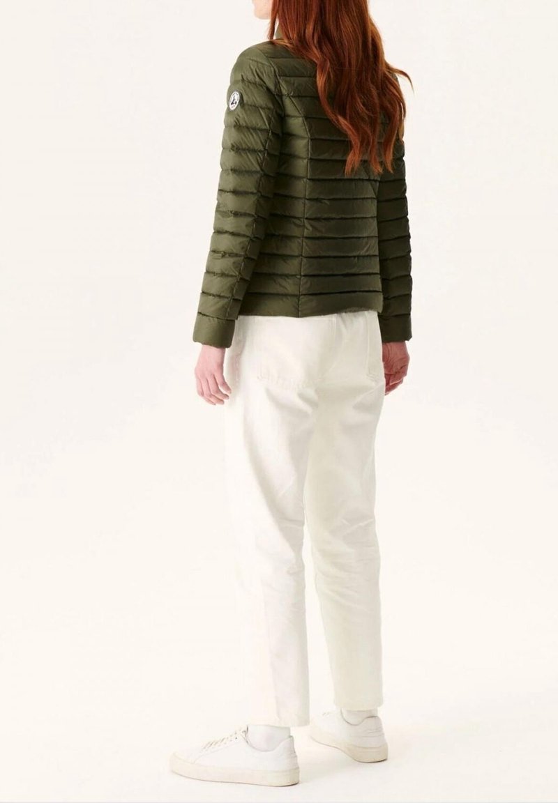 Femme aux longs cheveux roux portant une veste matelassée vert olive, un pantalon blanc et des baskets blanches, debout dos à nous.