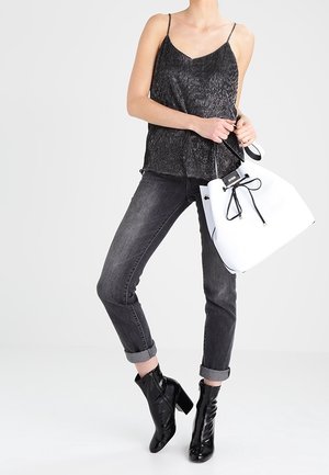 Vrouw in een zwarte mouwloze top, met opgestroopte donkergrijze jeans, zwarte enkellaarsjes met hak, en een grote witte handtas met zwarte trekkoord.