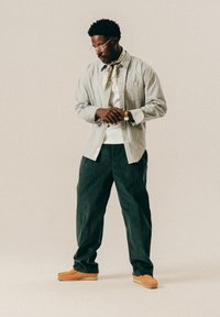 Mann mit Brille, beigem gestreiften Hemd, weißem T-Shirt, grüner Hose, hellbraunen Schuhen und einem gemusterten Halstuch, der auf seine Armbanduhr schaut.