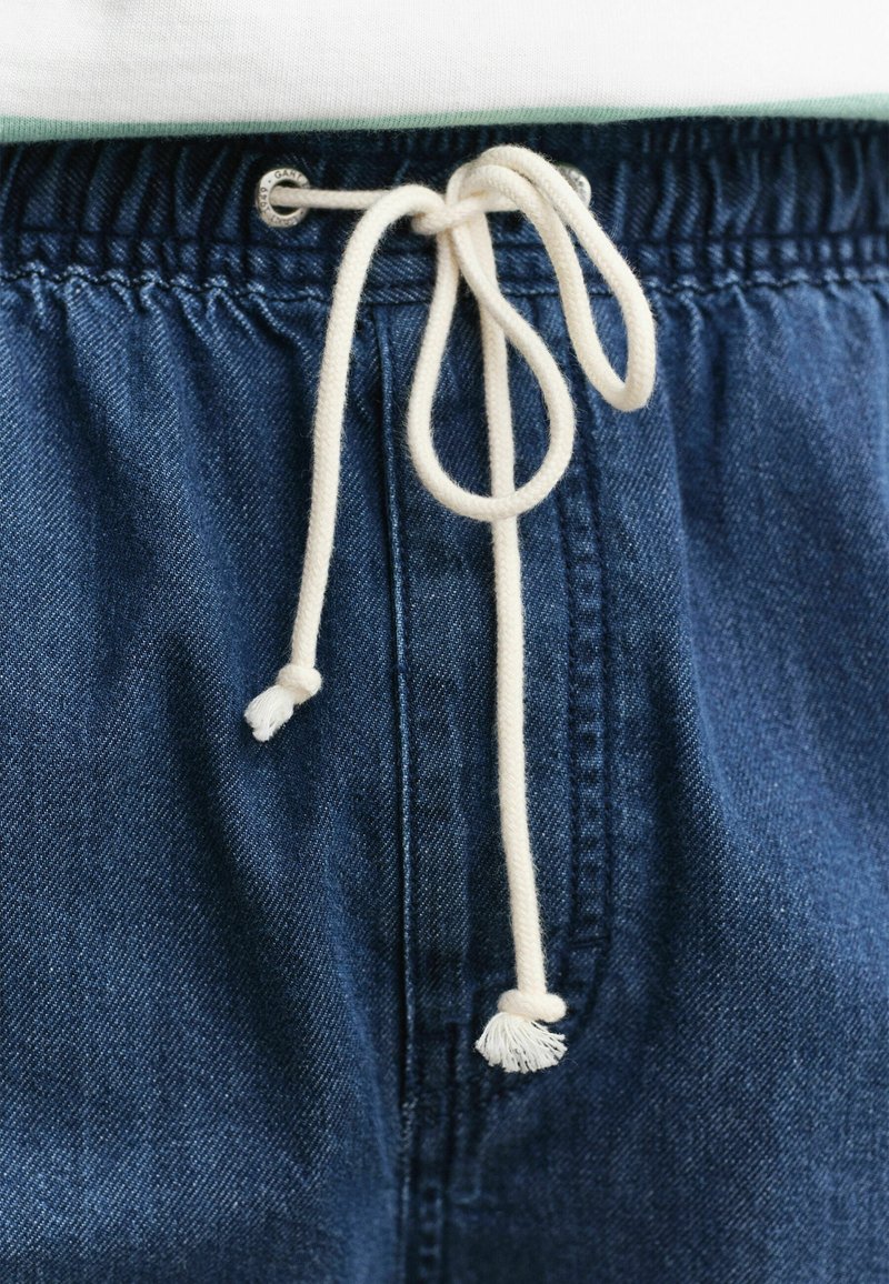 Primo piano di pantaloni in denim blu con coulisse bianca annodata a fiocco sulla vita elasticizzata sotto un indumento bianco.