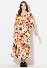 Blumenkleid in Midi-Länge mit kräftigen Mustern in Orange, Pink und Grün, kurzen Ärmeln, Knopfverschluss und einem taillierten Gürtel; kombiniert mit schwarzen Slippern.