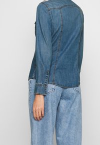 Denimjacka med medelblå tvätt, knäppning vid ärmsluten och en avslappnad passform. På baksidan finns sömdetaljer och en rundad nederkant.