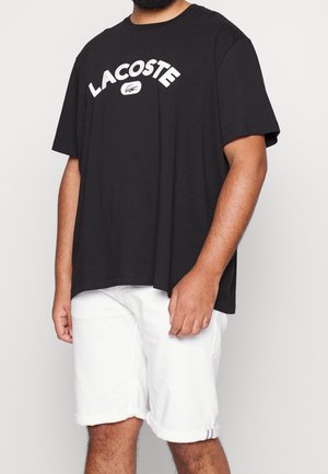 Mężczyzna w czarnym t-shircie Lacoste i białych krótkich spodenkach z podwiniętym brzegiem na jednolitym tle, pokazany od szyi do kolan.