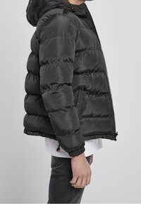 Homme portant une doudoune noire avec capuche, chemise blanche en dessous et pantalon sombre, debout de profil devant un fond gris.