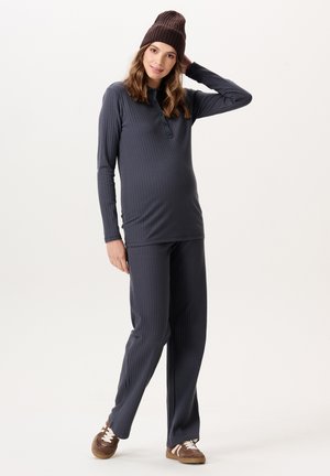 Zwangere vrouw draagt een geribd donkergrijs shirt met lange mouwen en een broek, een bruine muts en bruine sneakers, staand met één hand op haar hoofd.
