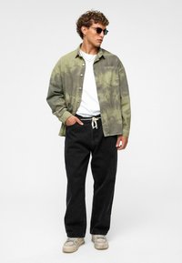 Olijfkleurige tie-dye shirtjack, witte t-shirt, zwarte broek met aansnoerkoord en lichtgekleurde sneakers. Draagt een zonnebril en minimale sieraden.