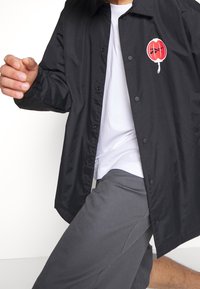 Svart knäppt jacka med en glänsande yta, med en röd cirkulär logotyp på vänster bröst och en krage.