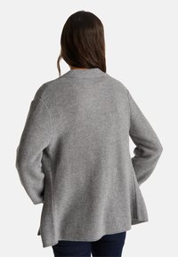 Suéter de punto gris con un corte holgado, escote redondo y mangas largas. Tejido texturizado con un detalle de dobladillo trasero ligeramente acampanado.