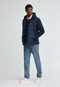 Marinblå pufferväst med huva, dragkedjestängning och sidofickor över en röd och grön randig tröja, kombinerad med ljusblå jeans och beige sneakers.