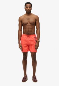 Pantaloni da bagno arancioni brillanti con vita regolabile, caratterizzati da un logo nero "DRY" sul lato sinistro. Indossati con scarpe marroni.