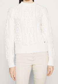 Vit stickad tröja med en mönstrad textur, rund halsringning och ribbad mudd. Ärmarna har upphöjda bollar som tillför detaljer.