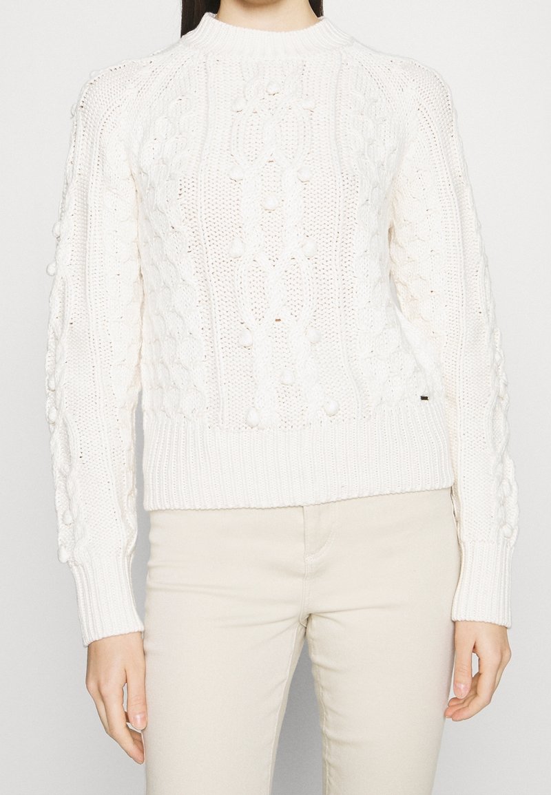 Vit stickad tröja med en mönstrad textur, rund halsringning och ribbad mudd. Ärmarna har upphöjda bollar som tillför detaljer.