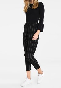 Schwarzes Langarmoberteil mit Glockenärmeln, kombiniert mit schwarz gestreiften, taillierten Hosen und weißen Sneakern. Der Stoff sieht glatt aus, mit minimaler Textur.