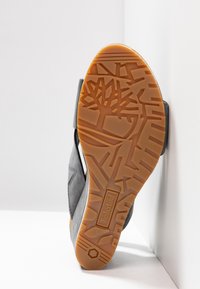 Semelle d'une chaussure Timberland grise avec une bande de roulement en caoutchouc beige arborant un logo d'arbre et des motifs géométriques, inclinée contre un mur blanc et un sol.