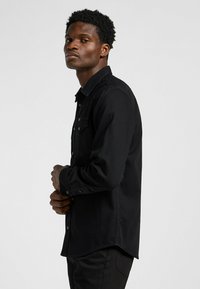 Camisa negra de manga larga con cuello abotonado, bolsillo en el pecho y dobladillo recto, hecha de una tela texturizada. Llevada con pantalones oscuros.