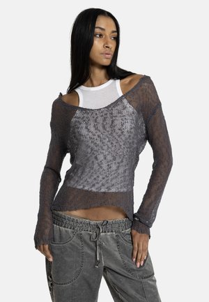 Femme portant un pull transparent gris en maille épaules dénudées par-dessus un débardeur blanc et un pantalon ample gris à cordon, debout avec une main dans la poche.