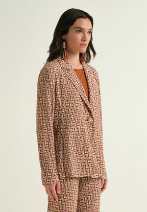 Frau trägt einen Blazer mit geometrischem Muster und passende Hose, orangefarbene Quastenohrringe und ein oranges Oberteil, stehend vor einem schlichten Hintergrund.