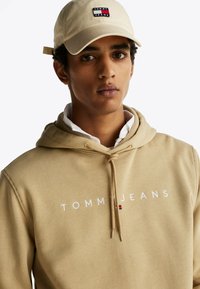 Beżowa bluza z kapturem z logo "TOMMY JEANS" w kolorze białym, z regulowanym sznurkiem i teksturowanym materiałem. Powyżej beżowa czapka z jasnym logo.