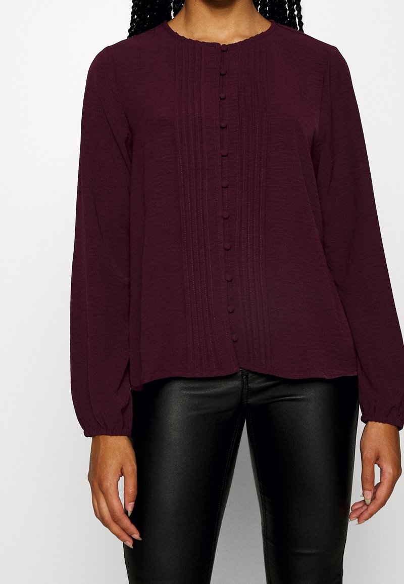 Femme portant un chemisier à manches longues violet foncé avec des boutons et des plis verticaux, associé à un pantalon en cuir noir.