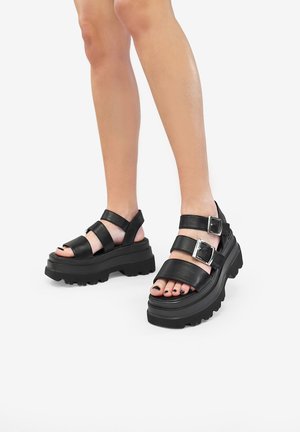 Black leather platform sandals with thick soles, featuring multiple straps and metallic buckles. Exposed toes with black nail polish visible.