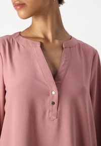 Rosa bluse med glatt stoff, V-hals med knappestolpe og subtile gullakenter. Enkel, avslappet design med kortermede ermer.