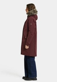 Bordeaux parka med en pelsforet hætte, der har en lige pasform, lynlås lukning og sidelommer. Båret over mørk denim og sorte støvler.