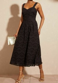 Žena v černých midi šatech s širokými ramínky, držící zlatou clutch kabelku a obutá ve zlatých sandálech na vysokém podpatku.