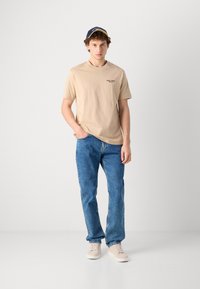 Beige bomulds-t-shirt med lille sort tekst, parret med blå denimjeans og lyse beige sneaks. Modellen bærer en marineblå kasket.