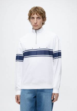 Jeune homme aux cheveux bouclés portant un sweat zippé blanc avec des rayures horizontales bleu marine et le texte « HUGO », associé à un jean bleu.