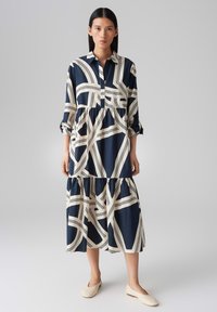 Button-up dress in navy and cream with abstract striped pattern. Features long sleeves, tiered hem, and lightweight fabric. Plain beige shoes.