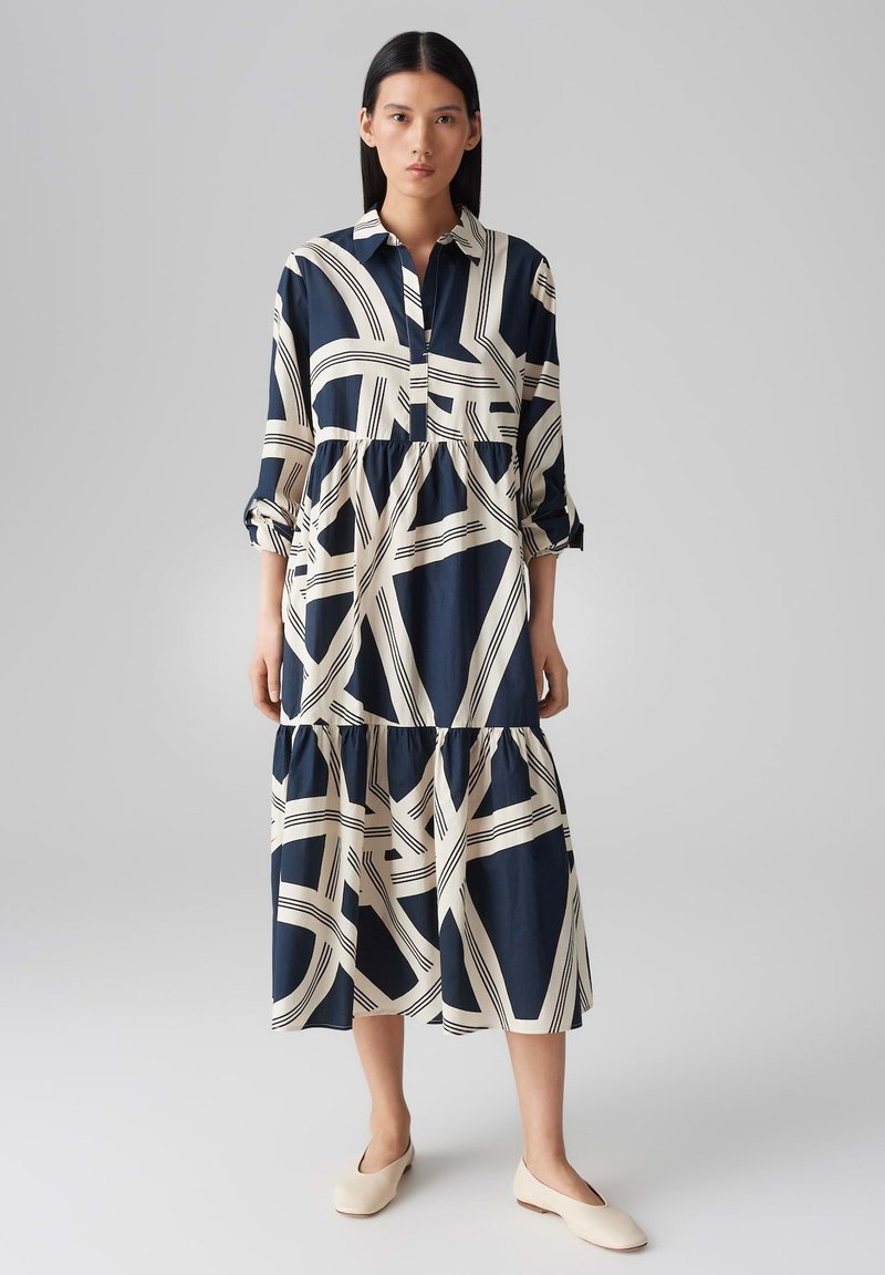 Button-up dress in navy and cream with abstract striped pattern. Features long sleeves, tiered hem, and lightweight fabric. Plain beige shoes.