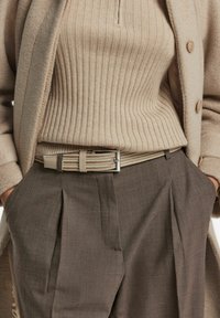 Vrouw in een beige geribbelde trui, ingestopt in een bruine plooibroek met een geweven beige riem en een beige jas met knopen, met de handen in de zakken.