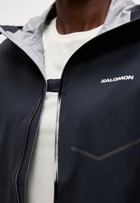 Κοντινό πλάνο ατόμου που φοράει μαύρο μπουφάν Salomon με γκρι εσωτερικό και λευκή διχτυωτή μπλούζα από μέσα.