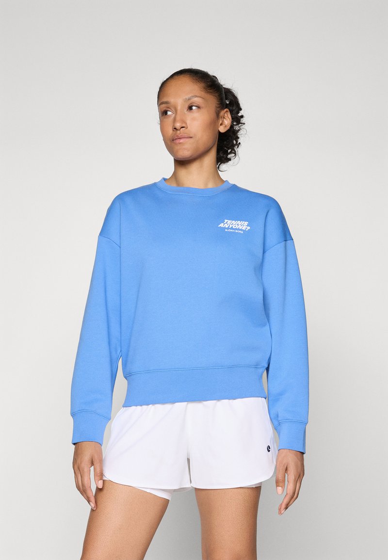 Sweatshirt azul feito de mistura de algodão, com gola redonda, mangas longas e um logótipo gráfico branco no lado esquerdo do peito. Estilizado com calções brancos.