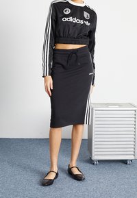 Person som bär en svart Adidas kortärmad tröja med långa ärmar och kjol med vita ränder, står bredvid en silverfärgad rullande förvaringslåda på en blå matta.