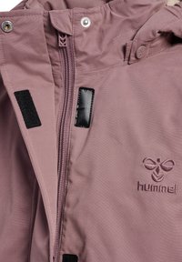 Gros plan de l'avant d'une veste mauve avec fermeture éclair, bouton-pression, bandes velcro et logo hummel brodé sur la poitrine.