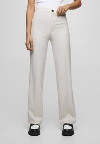 Pantalon à jambes larges en tissu beige clair avec une texture lisse. Dispose d'une fermeture par bouton et de poches frontales, associé à des chaussures à plateforme noires.