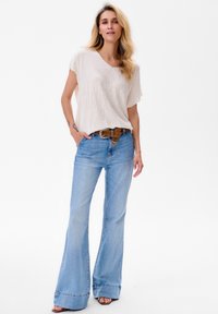 Cremige Bluse mit Struktur, kombiniert mit hellblauen, ausgestellten Denim-Jeans und einem braunen Gürtel mit gemusteter Schnalle. Schlichte Sandalen runden das Outfit ab.