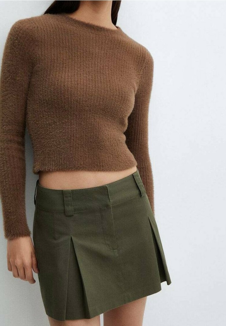 Femme portant un pull court marron à manches longues en tissu duveteux et une mini-jupe plissée vert olive sur un fond blanc uni.