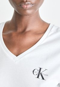 Tricou din bumbac alb, cu decolteu în V, având un mic logo negru "CK" și textul "Calvin Klein" în zona stângă a pieptului. Textură fină.