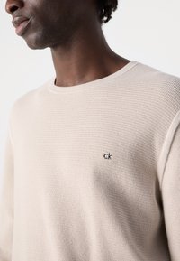 Beige textured knit sweater with a round neckline and subtle "CK" logo embroidered on the left side of the chest.